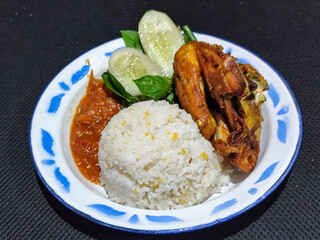 Lalapan ayam goreng. Crispy golden fried chicken dishes, with bright red chili sauce, fresh cucumber slices and a few basil leaves on a white enamel plate decorated with blue.