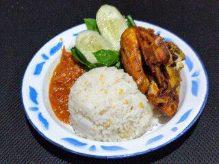 Lalapan ayam goreng. Crispy golden fried chicken dishes, with bright red chili sauce, fresh cucumber slices and a few basil leaves on a white enamel plate decorated with blue.