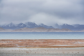 Grass and stone shore of Lake Karakul with blue water and mountain peaks in the background, cloudy...