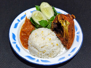 Lalapan ayam goreng. Crispy golden fried chicken dishes, with bright red chili sauce, fresh cucumber slices and a few basil leaves on a white enamel plate decorated with blue.