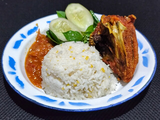 Lalapan ayam goreng. Crispy golden fried chicken dishes, with bright red chili sauce, fresh cucumber slices and a few basil leaves on a white enamel plate decorated with blue.