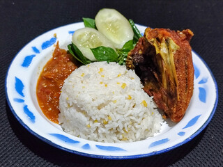 Lalapan ayam goreng. Crispy golden fried chicken dishes, with bright red chili sauce, fresh cucumber slices and a few basil leaves on a white enamel plate decorated with blue.