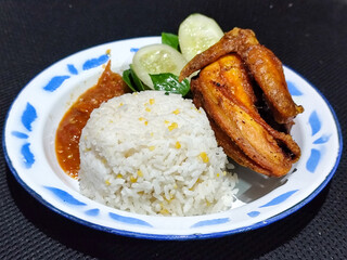Lalapan ayam goreng. Crispy golden fried chicken dishes, with bright red chili sauce, fresh cucumber slices and a few basil leaves on a white enamel plate decorated with blue.