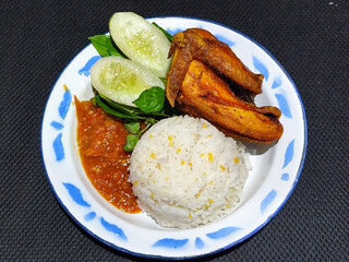 Lalapan ayam goreng. Crispy golden fried chicken dishes, with bright red chili sauce, fresh cucumber slices and a few basil leaves on a white enamel plate decorated with blue.