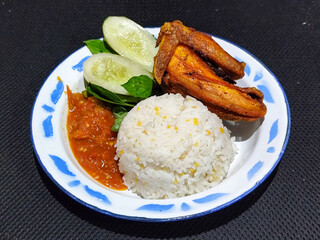 Lalapan ayam goreng. Crispy golden fried chicken dishes, with bright red chili sauce, fresh cucumber slices and a few basil leaves on a white enamel plate decorated with blue.