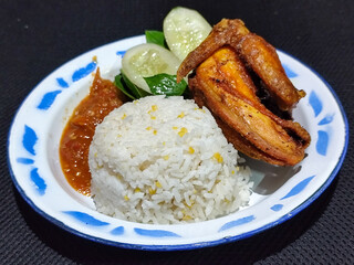 Lalapan ayam goreng. Crispy golden fried chicken dishes, with bright red chili sauce, fresh cucumber slices and a few basil leaves on a white enamel plate decorated with blue.