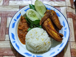 Lalapan ayam goreng. Crispy golden fried chicken dishes, with bright red chili sauce, fresh cucumber slices and a few basil leaves on a white enamel plate decorated with blue.