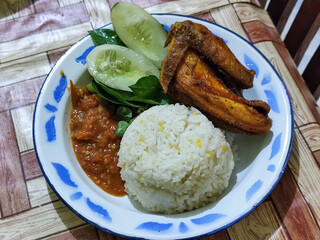 Lalapan ayam goreng. Crispy golden fried chicken dishes, with bright red chili sauce, fresh cucumber slices and a few basil leaves on a white enamel plate decorated with blue.