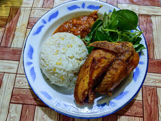 Lalapan ayam goreng. Crispy golden fried chicken dishes, with bright red chili sauce, fresh cucumber slices and a few basil leaves on a white enamel plate decorated with blue.