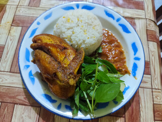Lalapan ayam goreng. Crispy golden fried chicken dishes, with bright red chili sauce, fresh cucumber slices and a few basil leaves on a white enamel plate decorated with blue.