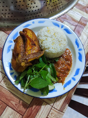 Lalapan ayam goreng. Crispy golden fried chicken dishes, with bright red chili sauce, fresh cucumber slices and a few basil leaves on a white enamel plate decorated with blue.