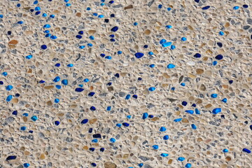 Close-up of a decorative beach pathway made of small natural pebbles embedded in concrete, with scattered pieces of shiny blue glass. The surface is part of a walking path or staircase near the sea, d