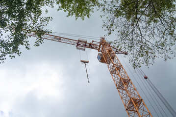Equipment for the construction and lifting of bulky goods with construction materials. A tower crane on a construction site against a rainy sky. A construction high-rise crane and leaves on a tree.