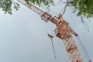 Equipment for the construction and lifting of bulky goods with construction materials. A tower crane on a construction site against a rainy sky. A construction high-rise crane and leaves on a tree.