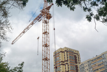 Equipment for the construction and lifting of bulky goods with construction materials. A tower crane on a construction site against a rainy sky. A construction high-rise crane and leaves on a tree.