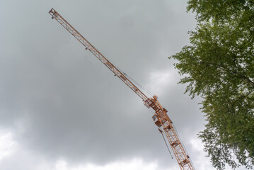 Equipment for the construction and lifting of bulky goods with construction materials. A tower crane on a construction site against a rainy sky. A construction high-rise crane and leaves on a tree.