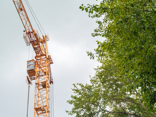 Equipment for the construction and lifting of bulky goods with construction materials. A tower crane on a construction site against a rainy sky. A construction high-rise crane and leaves on a tree.