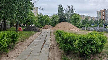 The sidewalk across a grassy lawn with acacia bushes growing on it is being repaired. Construction materials and piles of sand and asphalt crumbs lie nearby. Buildings stand around it. Overcast spring