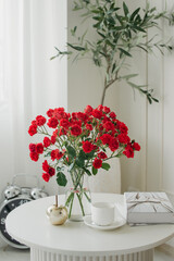 red roses in a vase stand on a white table next to a book and a cup of tea or coffee, in a white studio location or room decorated for an advertising shoot or photo shoot
