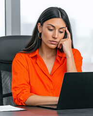 Young woman working on laptop in office wearing orange shirt with thoughtful expression