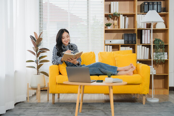 Reading on Yellow Sofa: An Asian woman is reading a book on yellow sofa while she put laptop computer on the small table in the living room.
