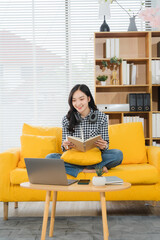 Cozy Reader: A cheerful woman sits comfortably on a vibrant yellow couch, engrossed in a book, laptop nearby, creating a perfect image of relaxation and study.