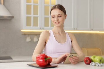 Woman using smartphone at white table with kitchen scale, healthy food and measuring tape indoors