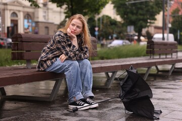Frustrated woman sitting near broken umbrella on bench outdoors