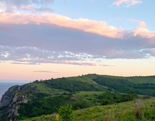 Obraz premium Lush green hills meet ocean under a pastel, cloud-strewn sky