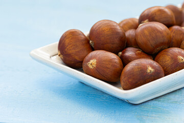Close-up of raw brown chestnuts piled on a white ceramic plate over blue wooden table, natural food ingredient and healthy snack concept.