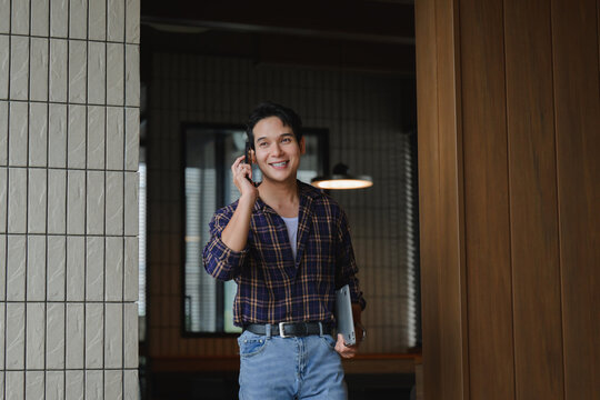 Young asian freelancer walking out of his office talking on the phone and holding a laptop - Powered by Adobe