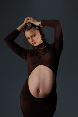 Beautiful pregnant young blonde in a brown dress with an open belly stands against a white wall....