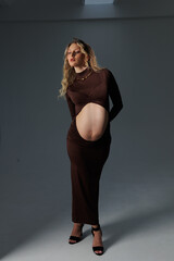 Beautiful pregnant young blonde in a brown dress with an open belly stands against a white wall....