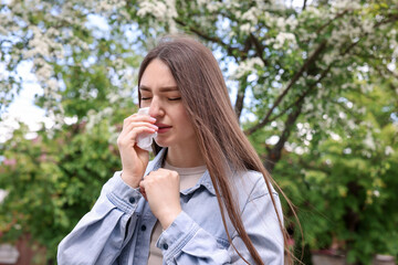 Woman with paper tissue suffering from seasonal pollen allergy near blossoming tree on spring day