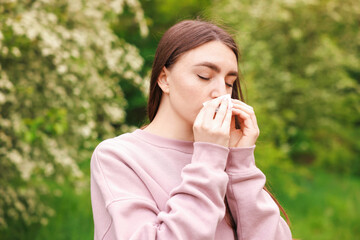 Woman with paper tissue suffering from seasonal pollen allergy near blossoming shrub on spring day