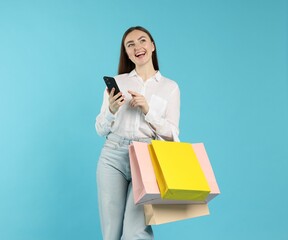 Positive woman with smartphone and shopping bags on light blue background