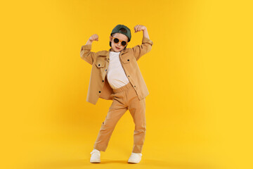 Cute little boy in sunglasses and baseball hat dancing on orange background