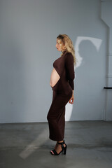 Beautiful pregnant young blonde in a brown dress with an open belly stands against a white wall....