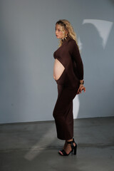 Beautiful pregnant young blonde in a brown dress with an open belly stands against a white wall....