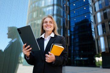 Confident businesswoman stands outside modern skyscrapers, holding tablet and notebooks, excited...