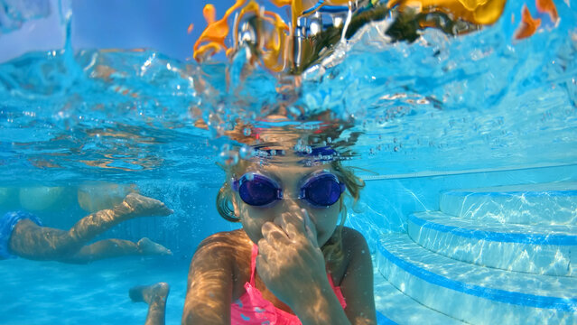 A young girl of caucasian ethnicity, wearing goggles and a pink swimsuit, swims underwater in a pool. The camera captures a close-up shot from below, emphasizing the joy and excitement of summer fun.