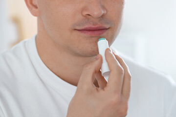 Young man applying lip balm onto his lips at home, closeup