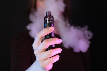 Woman using electronic smoking device in neon lights on black background, closeup
