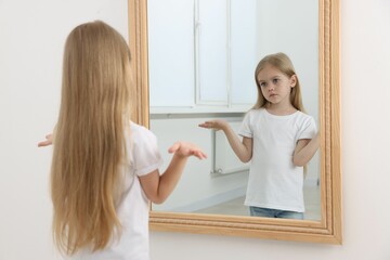 Upset little girl looking at herself in mirror indoors