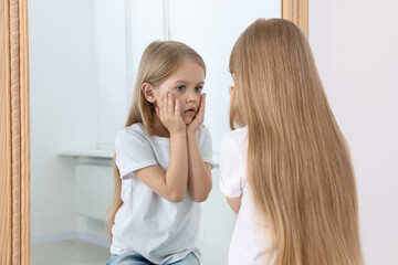 Upset little girl looking at herself in mirror indoors