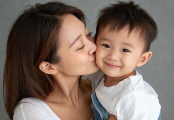 an Asian woman holding her son, dressed in blue jeans and a white shirt, kissing the boy on his forehead against a light gray background