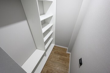 Fototapeta premium narrow, empty walk-in closet features white walls and shelves. The floor is tiled in a wood-like pattern. A wall switch is visible on the right
