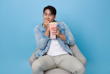 young man sitting chair looks visibly scared biting his nails while holding bucket of popcorn...