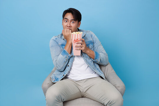 young man sitting chair looks visibly scared biting his nails while holding bucket of popcorn against isolated on blue background perfect for conveying suspense or fear.