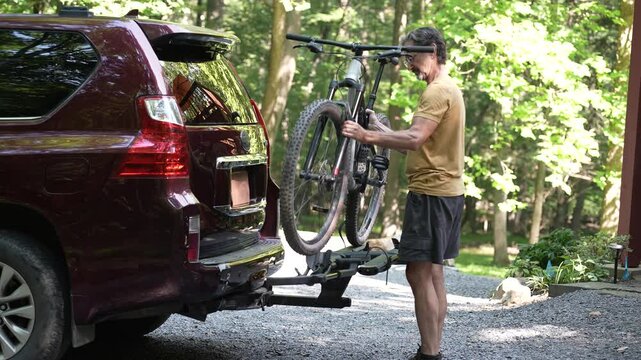 A person is securely placing a bike onto a hitch-mounted rack on the back of a car in a scenic wooded location, preparing for an outdoor biking adventure.
